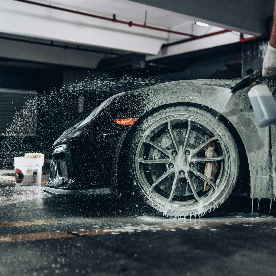 A person spraying soapy foam onto a black sports car in a garage